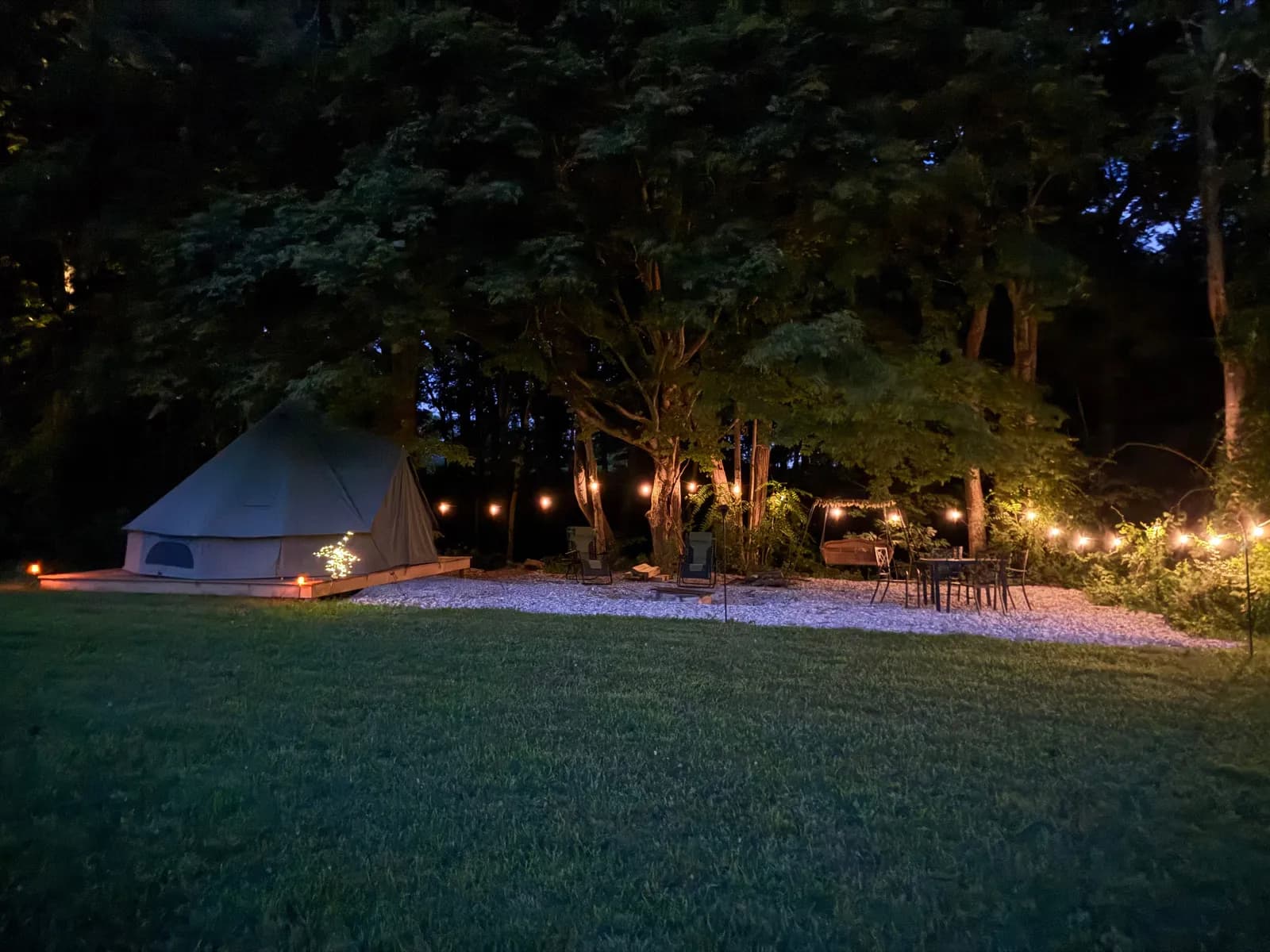 The Moonlight Meadow bell tent glowing on its deck at night, warm string lights strung through the surrounding trees, with zero-gravity loungers, a fire pit and a dining set arranged on the gravel pad beside the dark meadow.