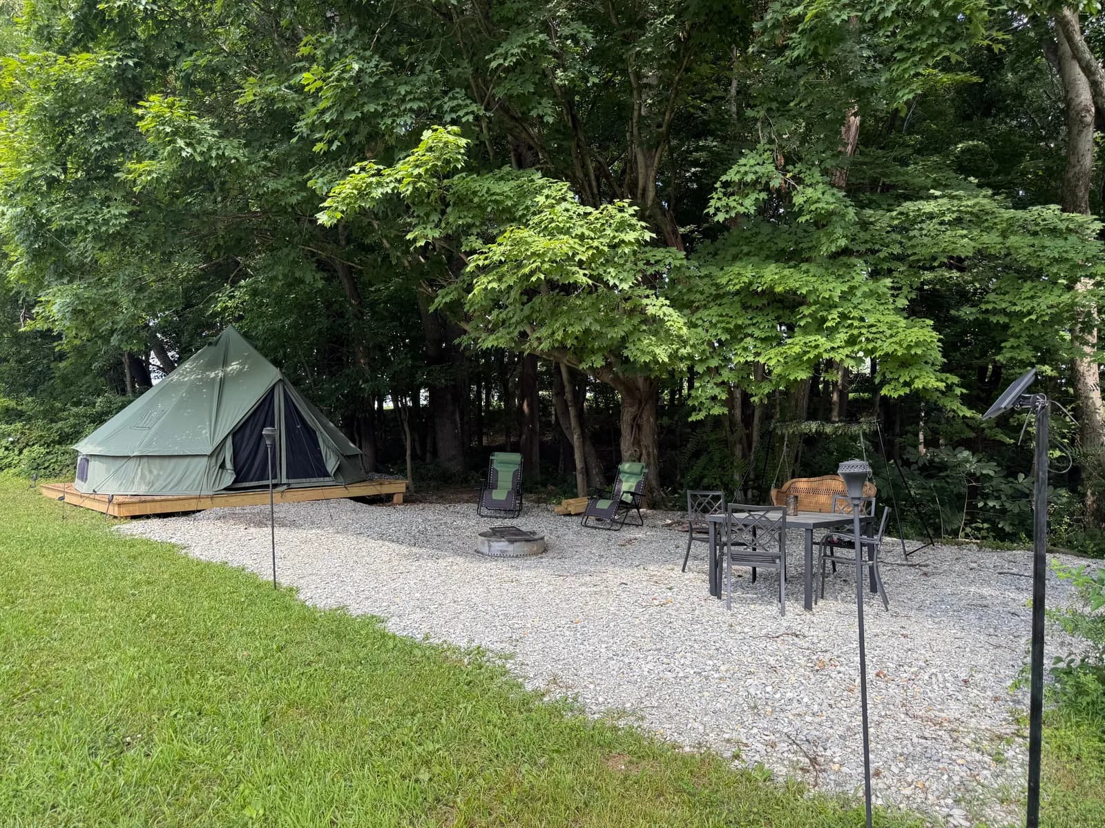 The green 16-foot bell tent on a wooden platform by day, set against leafy trees, with a fire pit ring, two green zero-gravity loungers, a metal dining table and chairs, and a hanging wicker porch swing on the gravel pad, the open meadow at the edge.