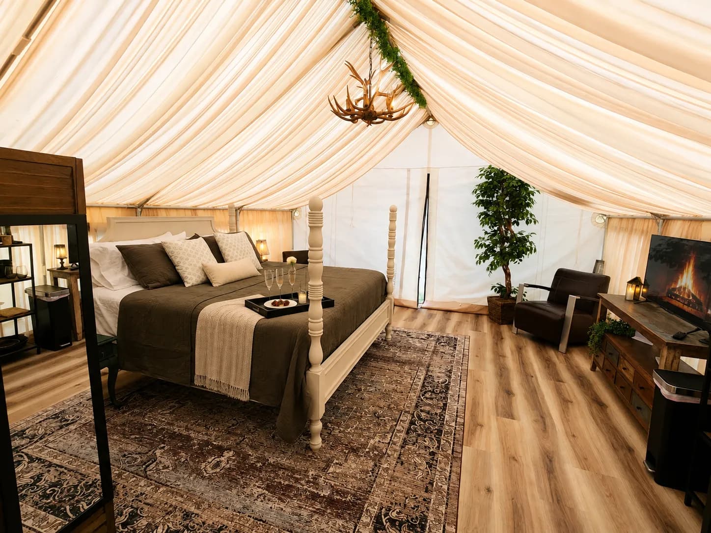 Inside The Velvet Buck: a four-poster bed with layered olive and cream linens under a draped canvas ceiling, an antler chandelier above, and champagne flutes and chocolate-covered strawberries on a tray at the foot of the bed.