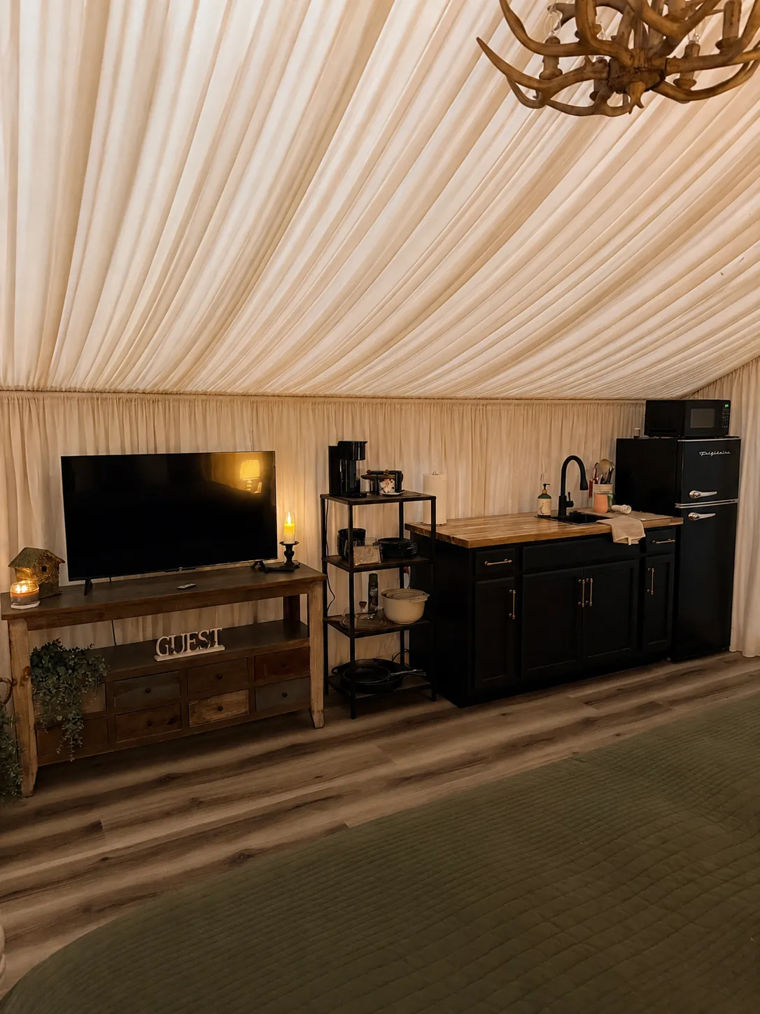 The Velvet Buck's kitchenette: a black cabinet island with butcher-block counter and sink, a mini fridge with freezer, a microwave, a coffee maker, stocked cookware and dishes, and a flat-screen TV on a wooden console.